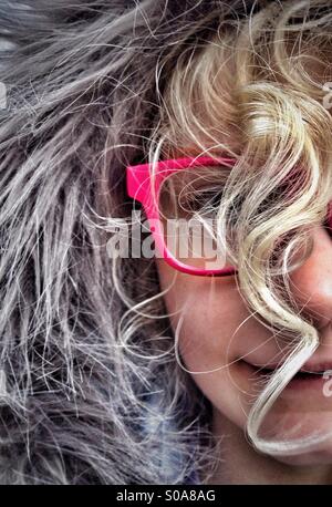 Jeune fille portant des lunettes rouges et capot furry avec visage masqué par des cheveux blonds bouclés Banque D'Images