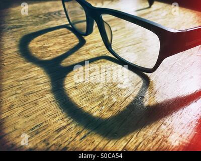 Une paire de lunettes de lecture d'un rebord épais assis sur un bureau en bois jette une ombre dans la lumière du soleil.