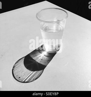 Le soleil qui brille dans un bécher d'eau en plastique jette une ombre sur une table blanche. Banque D'Images