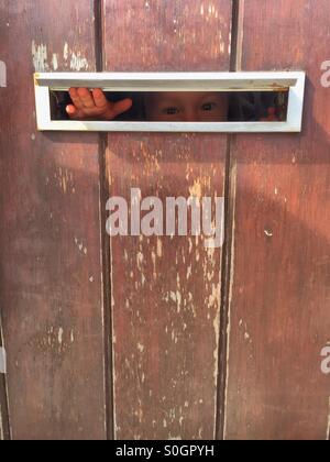 Peu peur enfant Peeking through la boîte aux lettres d'une porte. Banque D'Images