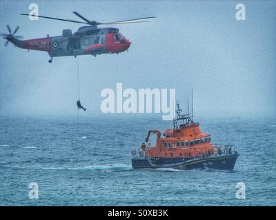 La Royal Navy et l'Air Sea Rescue helicopter descente d'un homme sur un treuil de sauvetage de la RNLI en attente sur un océan brumeux. Banque D'Images