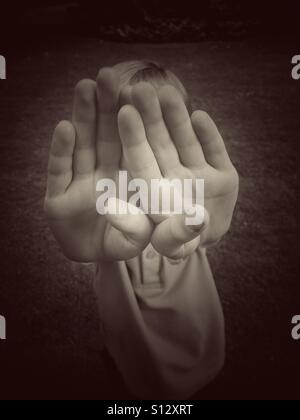 Un garçon de six ans, utilise ses deux mains pour couvrir ou protéger son visage. Le garçon est dans l'humeur ludique ou est-ce qu'il peur d'un pédophile attaque ? Une image pour illustrer les questions sociales. Banque D'Images