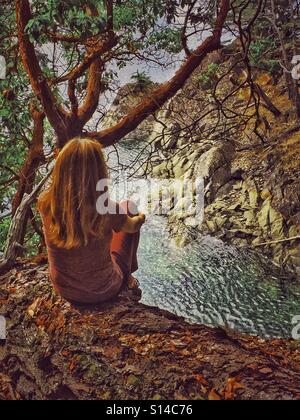 Une femme aux longs cheveux brun rouge donne à la vue sur l'eau à partir d'un point de vue élevé tout en étant assis sur un rondin en Colombie-Britannique, Canada. Banque D'Images