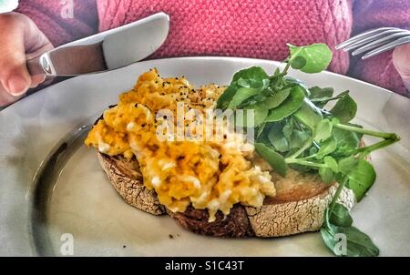 Le petit déjeuner ! Oeufs brouillés sur toast, femme et prêt à manger, avec couteau et fourchette en mains. Banque D'Images