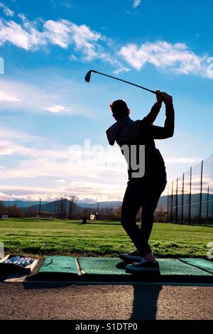 Jeune homme portant des vêtements de ville pratiquer son swing de golf à une gamme de conduite. Banque D'Images