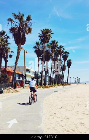 Journée de printemps ensoleillée dans le sud de la Californie. Un adolescent d'une bicyclette avec des palmiers et des maisons d'un côté et la plage de sable de l'autre. Banque D'Images