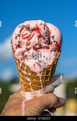 Photo verticale de la fonte de la crème glacée au chocolat cerise soit tenue dans un cornet gaufré contre un ciel bleu avec des nuages et des arbres en arrière-plan flou Banque D'Images