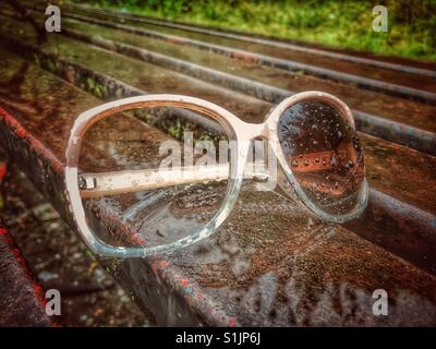 Paire de lunettes cassée avec un objectif et un bras sur un banc de parc sous la pluie Banque D'Images