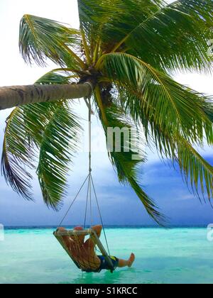 Jeune homme se détendre dans la pendaison swing du cocotier au fil de l'eau claire et profiter de la vue dans les Maldives. Banque D'Images