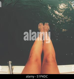 Les jambes bronzées de femme assise au bord d'un bateau avec les pieds immergés dans l'eau. S'est évanoui à modifier. Les carrés. Banque D'Images