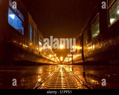 Vue en perspective de 2 trains stationnés pendant la nuit dans un dépôt, par une nuit humide. Prise de vue depuis la plate-forme. Banque D'Images