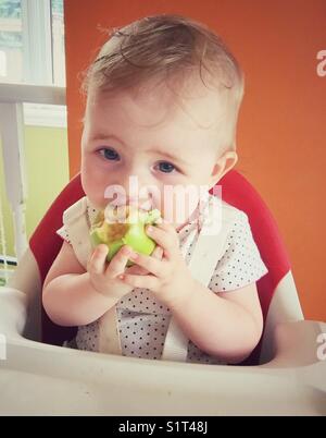 Bébé d'un an mangeant une pomme Granny Smith verte dans une chaise haute Banque D'Images