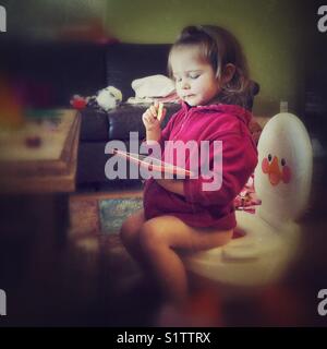 Formation toilettes bébé fille assise sur un pot dans une chambre familiale avec le comprimé dans une main et dans l'autre main craquelins Banque D'Images