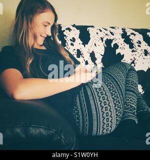 Image candide de 12 ans girl smiling at smartphone sur un canapé avec une couverture d'impression vache Holstein Banque D'Images