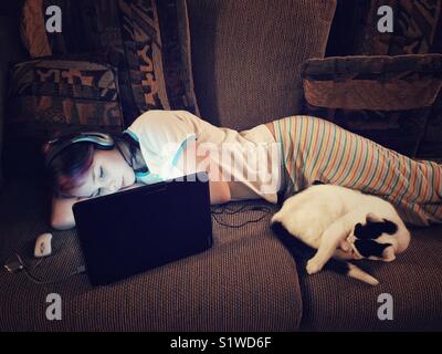 Girl laying on table en utilisant un ordinateur portable avec écouteurs et cat dormir à ses côtés Banque D'Images