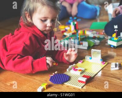 Bébé fille en rouge sweat à capuche portant sur plancher en bois LEGO jouer avec deux autres enfants partiellement en vue dans l'arrière-plan Banque D'Images