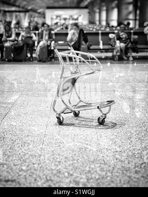 Chariot à bagages vide dans la zone de départ de l'aéroport avec des rangées de passagers assis à l'arrière-plan. Photo en monochrome Banque D'Images