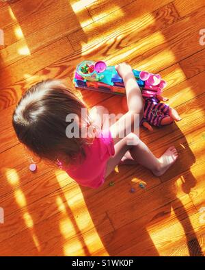 Bébé fille jouant avec les jouets sur plancher en bois avec la lumière du soleil faire un modèle de stores sur elle Banque D'Images