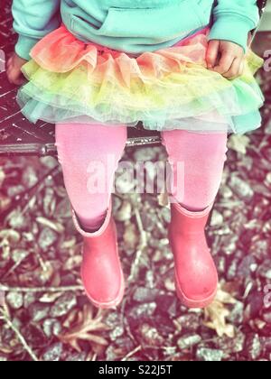 Petite fille assise sur une chaise à l'extérieur avec un tutu rose, collants et bottes de pluie rouge Banque D'Images