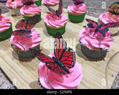 Cupcakes avec glaçage rose chaud et coloré papillons rouge et noir en haut affiche sur une planche à découper en bois. Banque D'Images