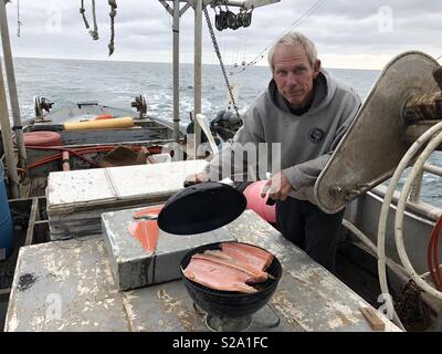 Handsome senior male pêcheur commercial prépare un saumon frais grillé sur le pont de son bateau de pêche vieux de 100 ans. Banque D'Images