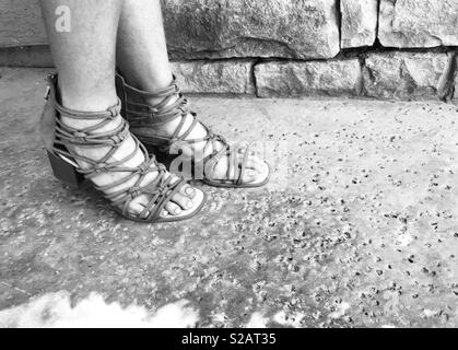 Photo en noir et blanc une femme en talon coincé brown Gladiator Sandals avec sangles , debout à l'extérieur sur le patio à côté d'un mur de pavés sur une journée d'été. Banque D'Images