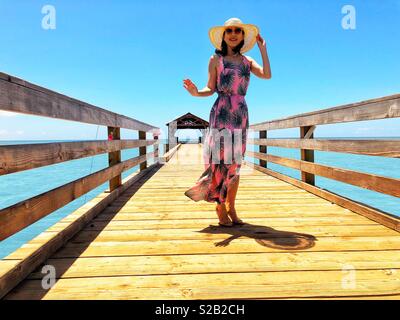 Belle jeune femme brune avec soutien figure, longue robe rose et vert floral rose, lunettes de soleil et hat smiling avec enthousiasme à bord d'une longue jetée de pêche en milieu tropical Kauai Hawaii Waimea. Banque D'Images