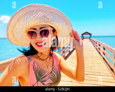 Belle jeune femme brune avec soutien figure, longue robe rose et vert floral rose, lunettes de soleil et hat smiling avec enthousiasme à bord d'une longue jetée de pêche en milieu tropical Kauai Hawaii Waimea. Banque D'Images