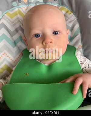 Bébé garçon aux cheveux blonds et yeux bleus, assis dans une chaise haute avec les pois verts sur le visage. Le port d'un dossard vert. Banque D'Images