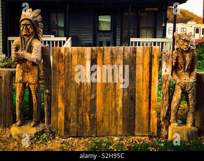 Statue en bois sculpté d'insoumis chef indien avec bras croisés et cowboy bourru avec les mains dans les poches de chaque côté du statif porte en bois en face de la maison Banque D'Images