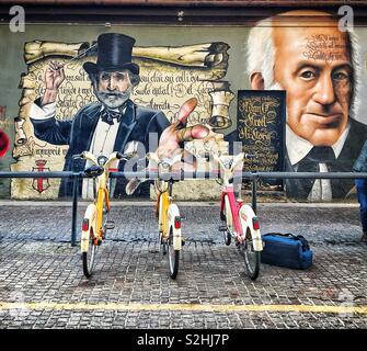 Trois vélos garés dans un rack à vélo sur le trottoir en face de l'art mural à côté d'un bâtiment à Milan, Italie. Un sac est sur le trottoir, à côté de l'un des vélos, appartenant à un jeune homme Banque D'Images