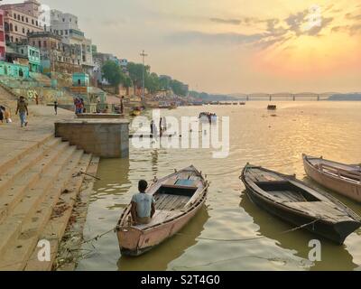 Vue d'un bateau l'homme dans son bateau pendant que les enfants jouent dans l'eau dans la distance au début du lever du soleil dans le Gange à Varanasi, Inde Banque D'Images