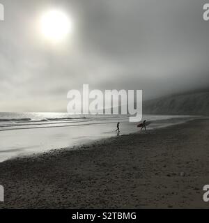 Deux surfeurs de sortir de l'eau silhouetté contre un soir lumineux soleil et brouillard marin lié à l'arrière-plan des falaises de craie à Compton Bay sur l'île de Wight Banque D'Images