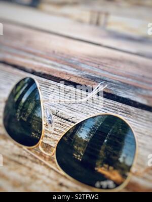 Lunettes de soleil aviateur assis sur une table de pique-nique en bois montrant le reflet des arbres dans la forêt sur l'objectif Banque D'Images