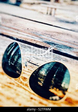 Lunettes de soleil aviateur assis sur une table de pique-nique en bois montrant le reflet des arbres dans la forêt sur l'objectif alors que le soleil brille sur eux Banque D'Images