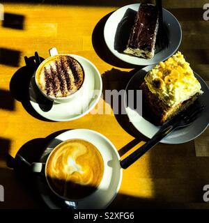 Cappuccino et Latte café en tasse et soucoupe avec des portions de gâteau sur les plaques latérales sur la table en bois éclairé ensoleillé avec des ombres projetées Banque D'Images