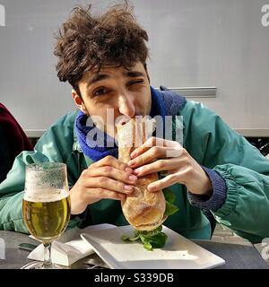 Jeune homme mangeant un sandwich de baguette fraîchement préparé. Banque D'Images