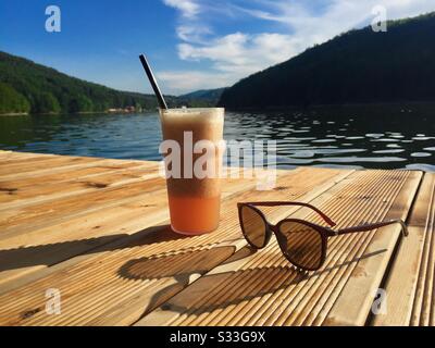 Verre de jus et paire de lunettes de soleil sur ponton en bois près du lac Banque D'Images