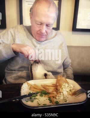 L'homme qui nourrit son poisson et ses chips, le chien est sous la table. Banque D'Images