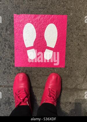 Image fantaisiste d'une femme debout à côté d'un pictogramme de distanciation sociale rose et blanc avec des chaussures roses assorties pendant la pandémie COVID-19, 2020, New York City, États-Unis Banque D'Images