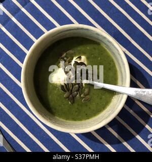 Un bol de soupe de légumes vert vif, recouvert de crème sure et de graines de citrouille, dans un bol blanc. Sur une table avec une nappe rayée bleue et blanche. Banque D'Images