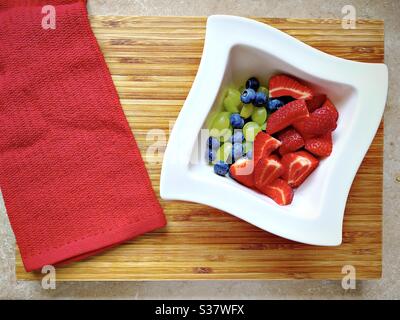 Vue rapprochée des baies fraîches de l'été dans un bol blanc sur une planche à découper en bois rustique. Préparer des fraises, des bleuets et des raisins sur le comptoir de cuisine. Banque D'Images