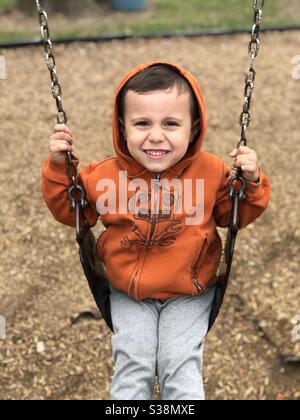 Un garçon de 4 ans sur une balançoire portant un veste à capuche tout en souriant Banque D'Images