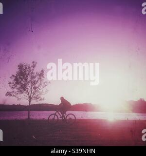 Photographie filtrée d'un homme qui traverse un parc à vélo au coucher du soleil avec un lac et un arbre Banque D'Images