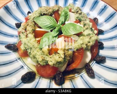 Gros plan d'une salade de caprese fraîche avec tomate, mozzarella, pesto de basilic, feuilles de basilic douces fraîches et raisins secs servis sur une assiette bleue et blanche Banque D'Images