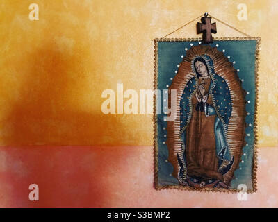 Une ombre est moulée dans un mur orange par une bannière de notre Dame de Guadalupe décorée d'une croix chrétienne dans une maison à Peña de Bernal, Queretaro, Mexique. Banque D'Images