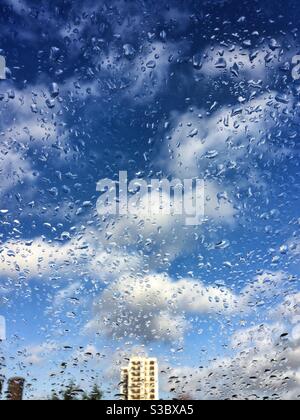 Ciel bleu et nuages blancs moelleux vus à travers une fenêtre mouiller avec des gouttes de pluie Banque D'Images