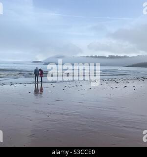 Couple sur la plage de Broadsands, Devon, Angleterre en hiver avec des nuages brumeux et de petites vagues en arrière-plan Banque D'Images