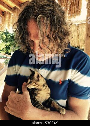 Homme hippy avec de longs cheveux dans un bleu et blanc t-shirt rayé dans un bar en plein air près de la piscine tenant un strain chaton brun et noir Banque D'Images