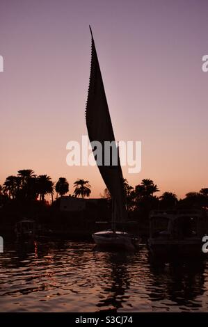 Beau grand felucca traditionnel fleuve Nil bateau navigue doucement sur le Nil au crépuscule avec le coucher du soleil jetant un rose, pêche, et violet doux lueur dans le ciel Banque D'Images